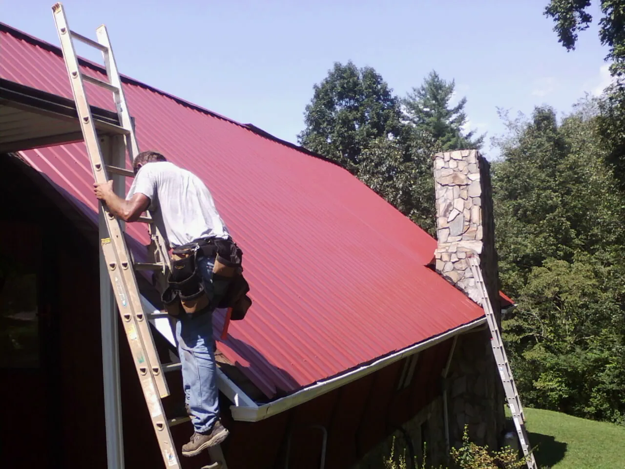 Roofer on a ladder beside a red metal roof and stone chimney — Soffit & Fascia Repair by Roofing Brothers in Rancho Cordova